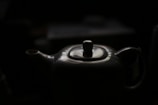 A porcelain teapot with a glossy surface is partially illuminated against a dark background. The spout and handle are visible, and the top lid has a round knob.