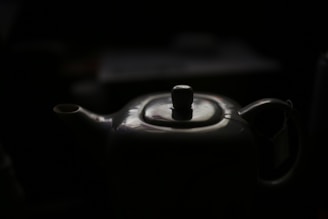 A porcelain teapot with a glossy surface is partially illuminated against a dark background. The spout and handle are visible, and the top lid has a round knob.