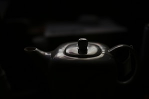 A porcelain teapot with a glossy surface is partially illuminated against a dark background. The spout and handle are visible, and the top lid has a round knob.