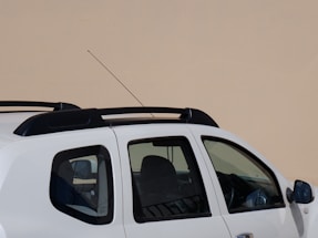 A partial view of a white SUV is visible, showing its side and part of the roof. The vehicle has tinted windows and a black roof rail system. The background is a plain beige wall, creating a stark contrast with the vehicle.