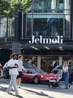 A busy urban scene in front of a department store named Jelmoli. Several people walk along the street, including a person with a stroller and two men talking. A red vintage sports car is parked on the street in front of storefronts displaying luxury brand names like Gucci and Dior.