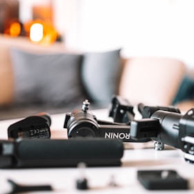 A close-up view of various camera equipment pieces, with a focus on a professional stabilizer or gimbal labeled 'Ronin'. The background features a blurred view of soft, warm lights and neutral-toned furniture, creating a cozy atmosphere.