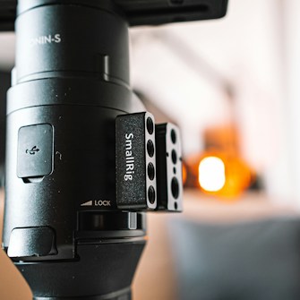 A close-up view of a piece of camera equipment marked with the label 'SmallRig'. The image features a black metallic component with several circular holes, attached to a larger cylindrical device with text indicating 'LOCK'. Some blurred bright orange and white lights are visible in the background, creating a soft and industrial atmosphere.