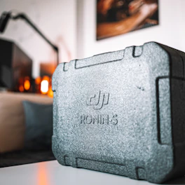 A close-up shot of a textured, grey carrying case with the brand name 'DJI Ronin-S' embossed on its surface. The background is slightly blurred, featuring indoor decor elements with warm lighting.