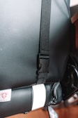 A close-up view of a black strap with a plastic buckle attached to a textured black leather surface. The bottom part of the image shows a section with stitched leather containing a white strip and a small logo. The buckle and strap suggest it may be part of a seat or bag.