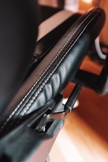Close-up view of a black leather office chair armrest with visible stitching and adjustment lever. Background is slightly blurred, showing some warm indoor lighting.