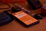 A smartphone with an orange screen displaying some text is lying on a wooden surface. Surrounding the phone are various objects including a blue fabric pouch, a smartwatch with a metallic strap, a camera lens cap labeled Nikon, and a black electronic device.