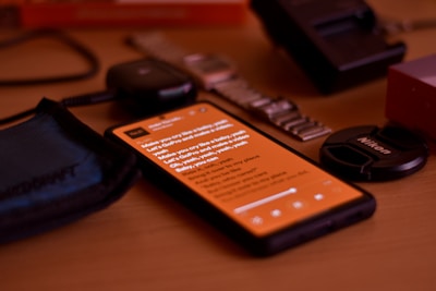 A smartphone with an orange screen displaying some text is lying on a wooden surface. Surrounding the phone are various objects including a blue fabric pouch, a smartwatch with a metallic strap, a camera lens cap labeled Nikon, and a black electronic device.