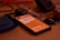 A smartphone with an orange screen displaying some text is lying on a wooden surface. Surrounding the phone are various objects including a blue fabric pouch, a smartwatch with a metallic strap, a camera lens cap labeled Nikon, and a black electronic device.