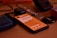 A smartphone with an orange screen displaying some text is lying on a wooden surface. Surrounding the phone are various objects including a blue fabric pouch, a smartwatch with a metallic strap, a camera lens cap labeled Nikon, and a black electronic device.