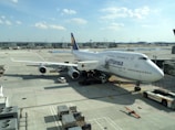 A large commercial airplane is parked at an airport gate. The aircraft is branded with the Lufthansa logo and is a Boeing 747 model. The surrounding area includes airport ground vehicles, luggage carts, and scattered airport equipment on the tarmac. In the background, the airport terminal is visible, along with a clear sky with scattered clouds.