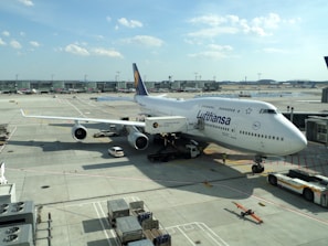 A large commercial airplane is parked at an airport gate. The aircraft is branded with the Lufthansa logo and is a Boeing 747 model. The surrounding area includes airport ground vehicles, luggage carts, and scattered airport equipment on the tarmac. In the background, the airport terminal is visible, along with a clear sky with scattered clouds.