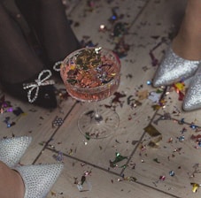 A glass filled with a pink beverage and adorned with colorful confetti on top is placed on a floor covered with more confetti. Two pairs of sparkly high heels are visible in the scene, suggesting a festive or celebratory atmosphere.