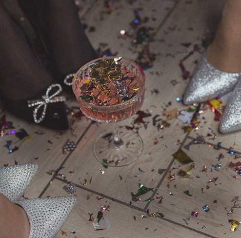 A glass filled with a pink beverage and adorned with colorful confetti on top is placed on a floor covered with more confetti. Two pairs of sparkly high heels are visible in the scene, suggesting a festive or celebratory atmosphere.