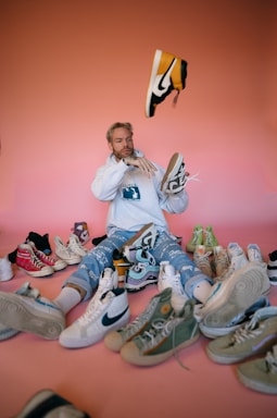 A person with blonde hair, wearing a light-colored hoodie and blue patterned pants, is surrounded by numerous pairs of sneakers on a gradient pink background. The person is holding one sneaker in each hand and tossing a yellow and black sneaker into the air.