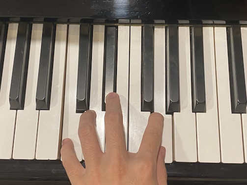 A close-up view of a person's hand playing a piano, with fingers pressing down on multiple keys. The image focuses on the piano keys, showcasing both the black and white keys in a clear and detailed manner.