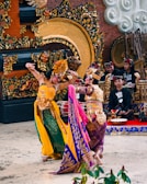 Two dancers in elaborate traditional costumes perform a cultural dance in front of a beautifully decorated ceremonial setting. The background features intricate carvings and musical instruments, with musicians wearing matching outfits sitting behind them. The dancers' costumes are colorful and ornate, showcasing detailed patterns and accessories.