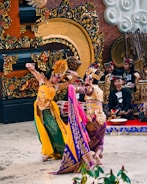 Two dancers in elaborate traditional costumes perform a cultural dance in front of a beautifully decorated ceremonial setting. The background features intricate carvings and musical instruments, with musicians wearing matching outfits sitting behind them. The dancers' costumes are colorful and ornate, showcasing detailed patterns and accessories.
