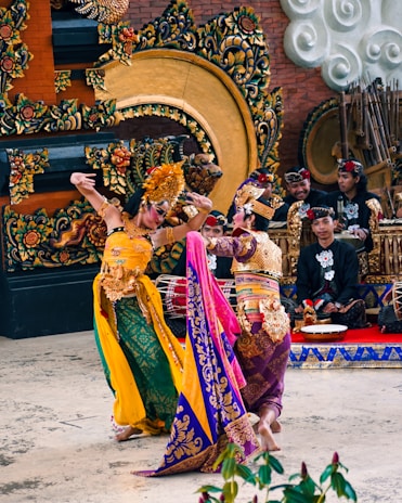 Two dancers in elaborate traditional costumes perform a cultural dance in front of a beautifully decorated ceremonial setting. The background features intricate carvings and musical instruments, with musicians wearing matching outfits sitting behind them. The dancers' costumes are colorful and ornate, showcasing detailed patterns and accessories.