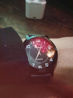 A wristwatch with a black strap and round black face displays white numerals and hands. The watch face reflects colorful bokeh lights. Behind the watch, there is a blurry, white rectangular object possibly resembling a small device against a dark brown wooden surface.
