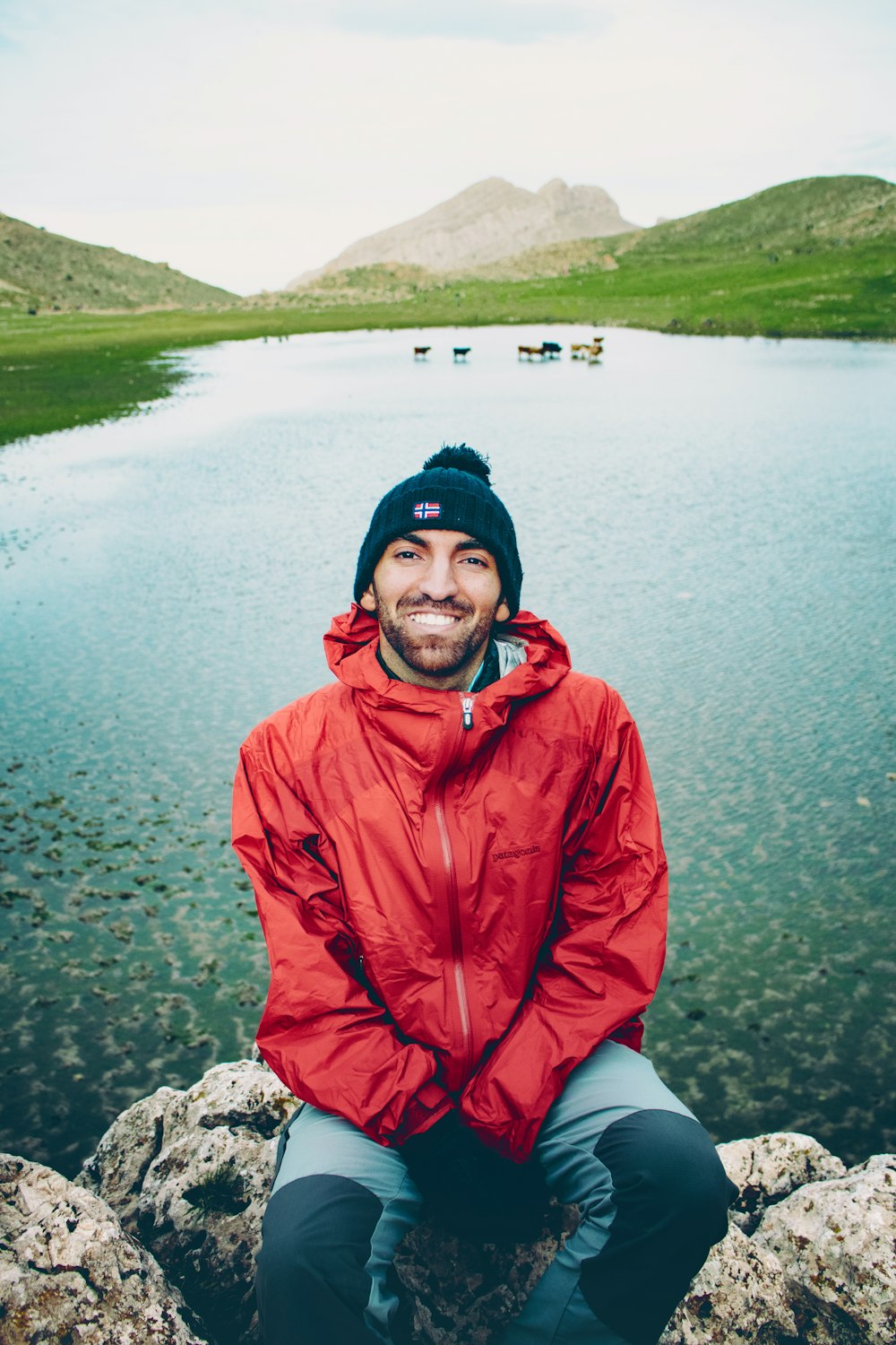 a man in a red jacket sitting on a rock by a lake