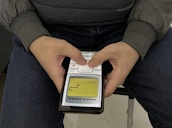 A person is holding an old, classic mobile phone with two hands, playing the Snake game. The person is wearing a gray jacket and sitting on a chair with blue jeans. The screen of the mobile phone displays the Snake game in progress with a yellow and black interface.