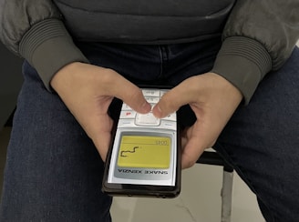 A person is holding an old, classic mobile phone with two hands, playing the Snake game. The person is wearing a gray jacket and sitting on a chair with blue jeans. The screen of the mobile phone displays the Snake game in progress with a yellow and black interface.