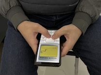 A person is holding an old, classic mobile phone with two hands, playing the Snake game. The person is wearing a gray jacket and sitting on a chair with blue jeans. The screen of the mobile phone displays the Snake game in progress with a yellow and black interface.