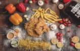 A flat lay of various food items on a rustic wooden surface. The arrangement includes crispy fried chicken, crinkle-cut fries, yellow and red bell peppers, cherry tomatoes, mushrooms, garlic cloves, and lime halves. There are also two small bowls of dipping sauces, dried pasta noodles, and a glass container with the words 'Beer Chicks' written on it. White flour is scattered across the scene, adding a rustic touch.