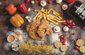 A flat lay of various food items on a rustic wooden surface. The arrangement includes crispy fried chicken, crinkle-cut fries, yellow and red bell peppers, cherry tomatoes, mushrooms, garlic cloves, and lime halves. There are also two small bowls of dipping sauces, dried pasta noodles, and a glass container with the words 'Beer Chicks' written on it. White flour is scattered across the scene, adding a rustic touch.