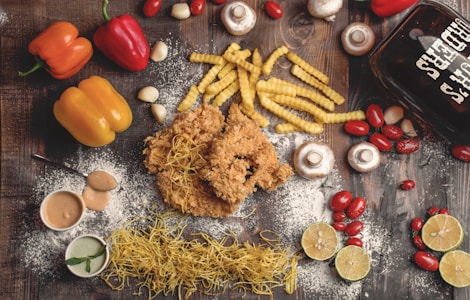 A flat lay of various food items on a rustic wooden surface. The arrangement includes crispy fried chicken, crinkle-cut fries, yellow and red bell peppers, cherry tomatoes, mushrooms, garlic cloves, and lime halves. There are also two small bowls of dipping sauces, dried pasta noodles, and a glass container with the words 'Beer Chicks' written on it. White flour is scattered across the scene, adding a rustic touch.