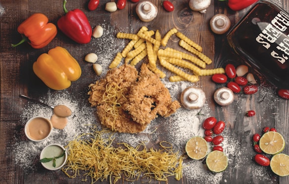 A flat lay of various food items on a rustic wooden surface. The arrangement includes crispy fried chicken, crinkle-cut fries, yellow and red bell peppers, cherry tomatoes, mushrooms, garlic cloves, and lime halves. There are also two small bowls of dipping sauces, dried pasta noodles, and a glass container with the words 'Beer Chicks' written on it. White flour is scattered across the scene, adding a rustic touch.