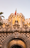 A historic castle facade featuring intricate stonework and decorative elements is topped with a national flag. The structure includes traditional crenellations and ornate sculptures, conveying architectural grandeur.