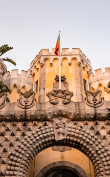 A historic castle facade featuring intricate stonework and decorative elements is topped with a national flag. The structure includes traditional crenellations and ornate sculptures, conveying architectural grandeur.