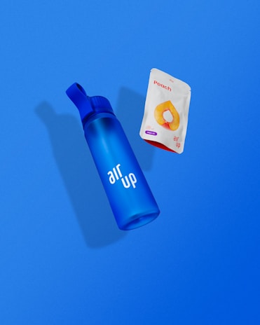 A blue bottle with the words 'air up' printed on it is positioned next to a packet labeled 'Peach' which features an image of a peach slice. Both items are set against a solid blue background.