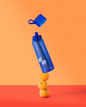 A blue water bottle with 'air up' written on it is balancing on top of three stacked oranges. The cap is hovering above the bottle as if it is thrown or unscrewed. The background is a vibrant orange with a red surface at the bottom.