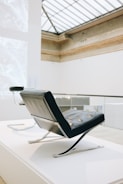 A modern chair with a sleek, angular design sits in an elegant, well-lit interior space with high ceilings and a large skylight. The chair features a combination of leather and metal elements, exuding a minimalist and sophisticated aesthetic.