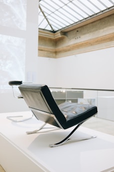 A modern chair with a sleek, angular design sits in an elegant, well-lit interior space with high ceilings and a large skylight. The chair features a combination of leather and metal elements, exuding a minimalist and sophisticated aesthetic.