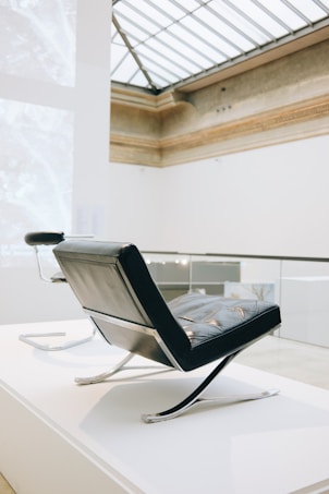 A modern chair with a sleek, angular design sits in an elegant, well-lit interior space with high ceilings and a large skylight. The chair features a combination of leather and metal elements, exuding a minimalist and sophisticated aesthetic.