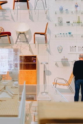 An array of modern chairs is displayed on transparent shelves in front of a pegboard wall. Various small objects such as vases, cameras, and utensils can be seen on the wall shelves. A person is standing in front of a wooden and chrome chair. The space has an organized, showroom-like atmosphere with a focus on design aesthetics.