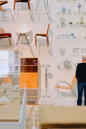 An array of modern chairs is displayed on transparent shelves in front of a pegboard wall. Various small objects such as vases, cameras, and utensils can be seen on the wall shelves. A person is standing in front of a wooden and chrome chair. The space has an organized, showroom-like atmosphere with a focus on design aesthetics.