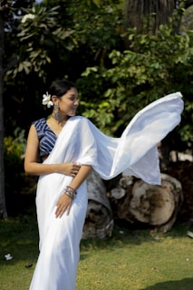A woman wearing a white sari with a blue blouse, adorned with large earrings and bracelets, stands outdoors. She has a flower in her hair and holds part of the sari that flows gracefully beside her. The background is lush with green foliage, and there are large, weathered logs on the ground.