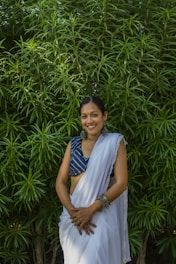 A woman is standing in front of lush green foliage, wearing a traditional white sari paired with a blue blouse. She has dark hair styled neatly and is adorned with jewelry including earrings and bangles. Her hands are clasped in front of her, and she is smiling warmly.