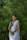 A woman is standing in front of lush green foliage, wearing a traditional white sari paired with a blue blouse. She has dark hair styled neatly and is adorned with jewelry including earrings and bangles. Her hands are clasped in front of her, and she is smiling warmly.