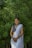 A woman is standing in front of lush green foliage, wearing a traditional white sari paired with a blue blouse. She has dark hair styled neatly and is adorned with jewelry including earrings and bangles. Her hands are clasped in front of her, and she is smiling warmly.