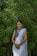 A woman is standing in front of lush green foliage, wearing a traditional white sari paired with a blue blouse. She has dark hair styled neatly and is adorned with jewelry including earrings and bangles. Her hands are clasped in front of her, and she is smiling warmly.