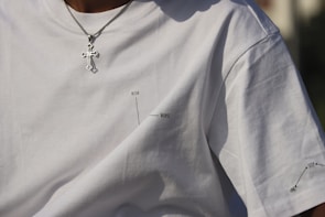 A close-up of a person wearing a plain white t-shirt featuring minimal black text with diagrams indicating 'better' and 'worse'. The person is also wearing a silver chain necklace with a cross pendant.