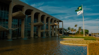 A modern architectural building with stone arches and distinctive water features stands beside a large reflective pool. Palm trees and greenery align the pathway, giving a tropical feel. The national flag waves on a pole against a backdrop of a clear blue sky.