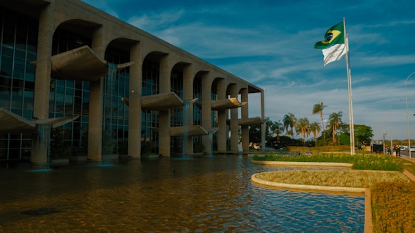 A modern architectural building with stone arches and distinctive water features stands beside a large reflective pool. Palm trees and greenery align the pathway, giving a tropical feel. The national flag waves on a pole against a backdrop of a clear blue sky.
