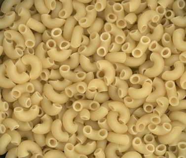 A close-up view of uncooked elbow macaroni pasta, showcasing a variety of small, curved tubes with hollow centers. The pasta pieces are uniformly yellowish in color, slightly arranged in a scattered pattern.